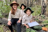 A teacher wearing a broadbrimmed hat sits on a log next to two students. All three are wearing khaki school shirts