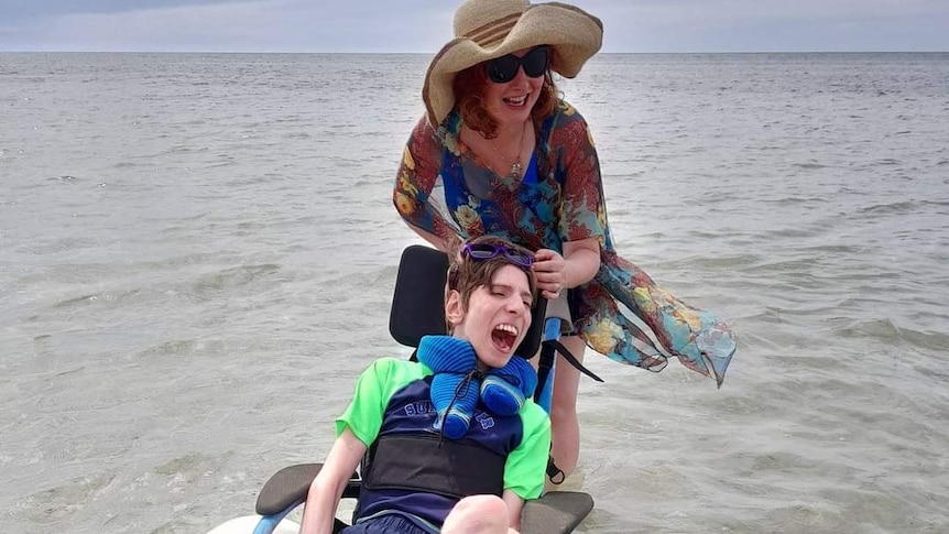 A smiling woman pushes her smiling son, who is in an aquatic wheelchair designed for the beach