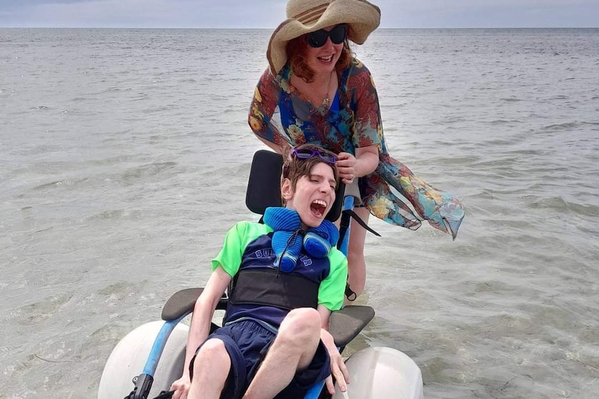 A smiling woman pushes her smiling son, who is in an aquatic wheelchair designed for the beach