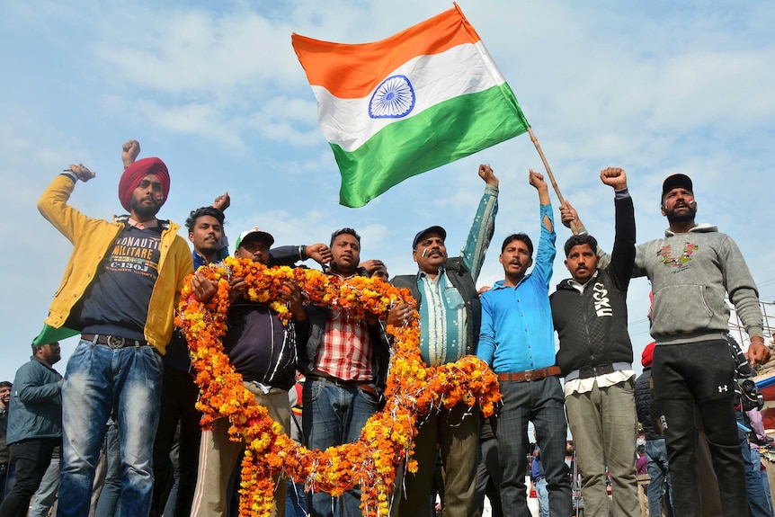 A line of men in a crowd outside raise one arm while also holding a large flower garland and Indian flag.