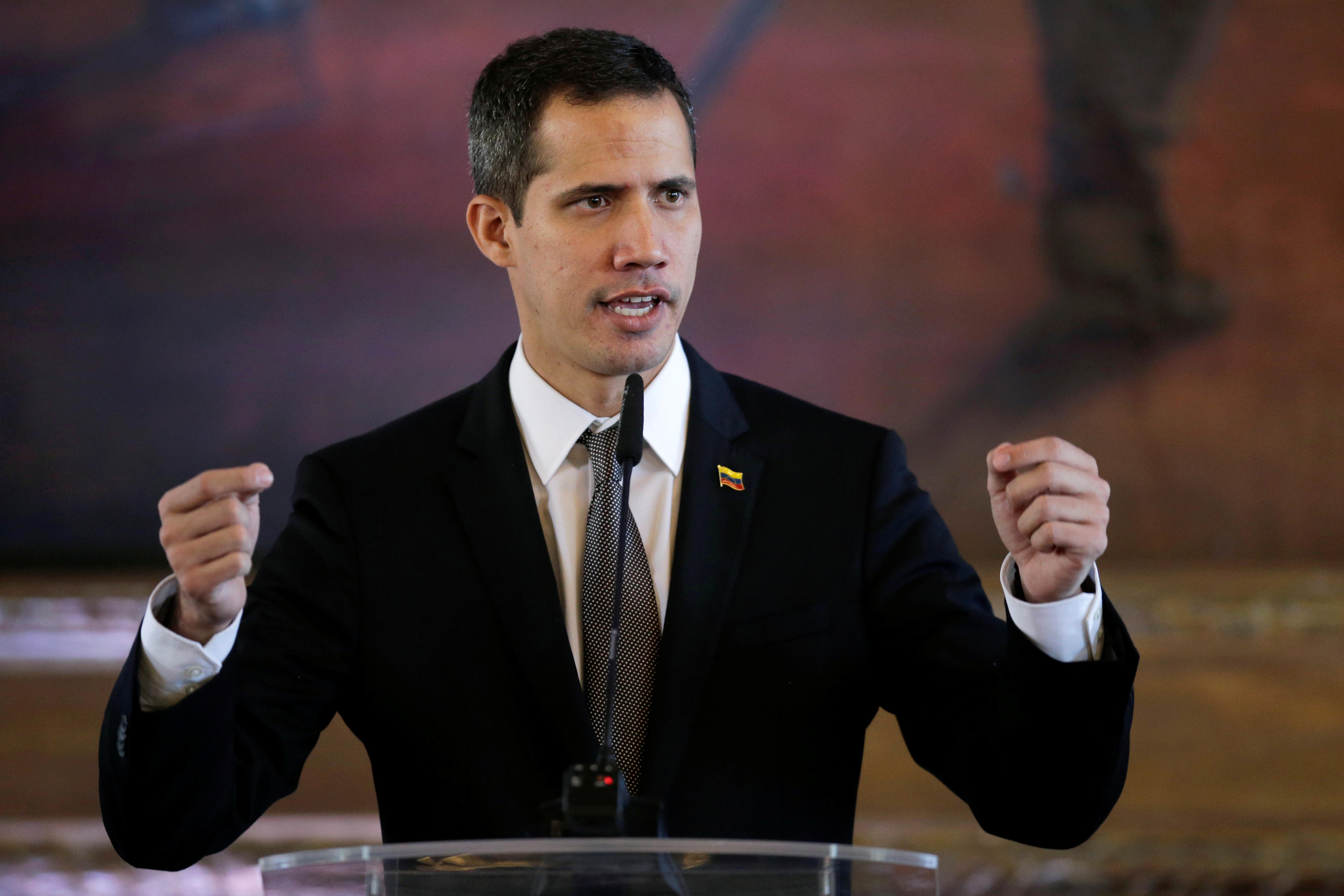 Juan Guaido in a black suit and tie speaking into a microphone while gesturing with two raised fists.