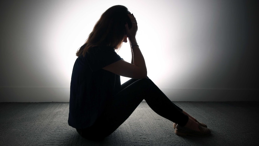 A woman silhouetted against a white wall sits with her head in her hands.