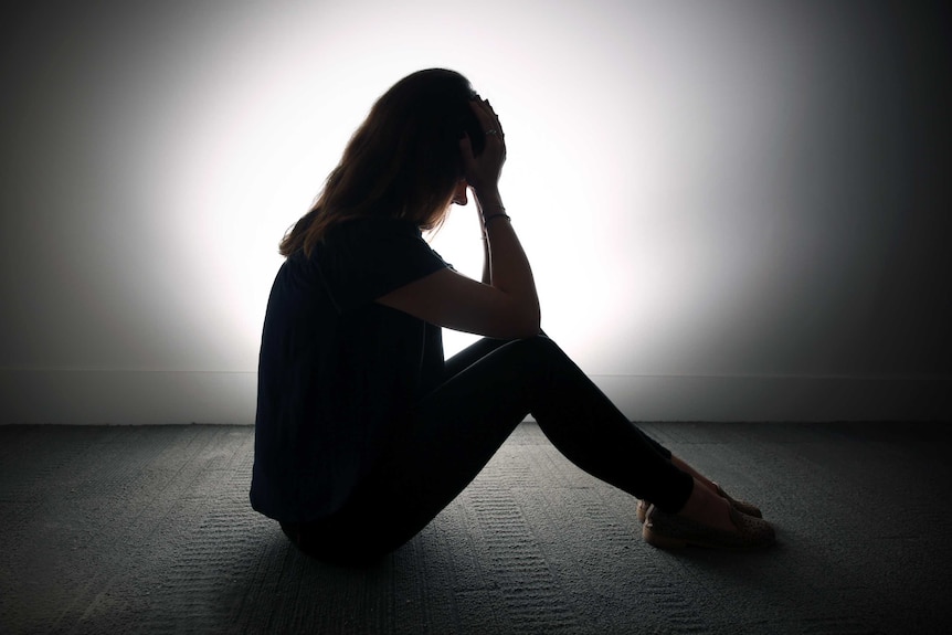 A silhouetted woman sits on the floor with her head in her hands.