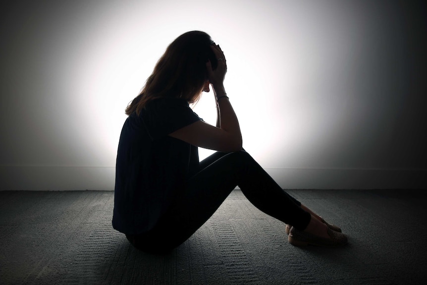 A woman silhouetted against a white wall sits with her head in her hands.