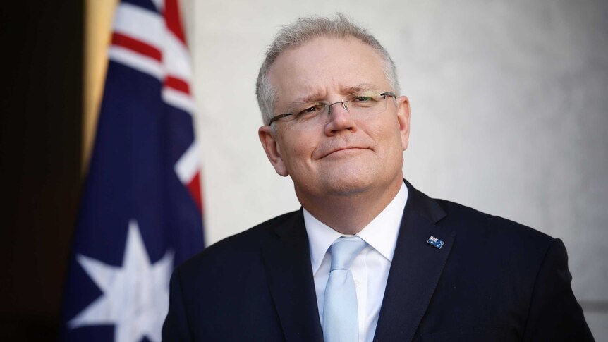 Morrison is standing in front on an Australian flag, looking into the distance smiling.
