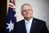 Morrison is standing in front on an Australian flag, looking into the distance smiling.