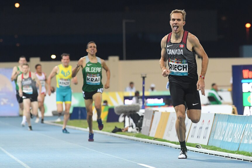 A man wearing black and red runs on a track poking his tongue out