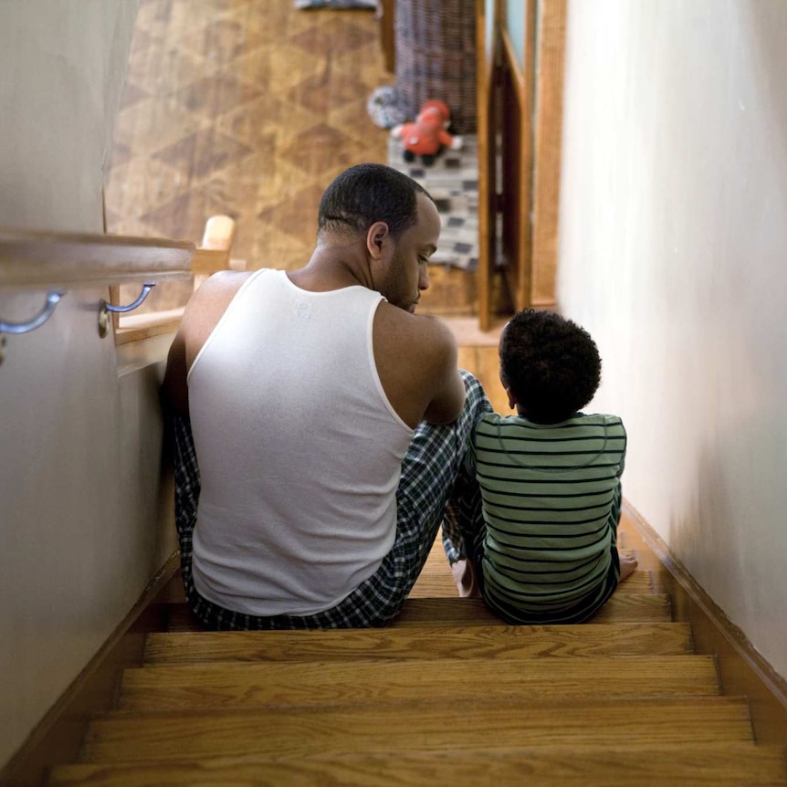 A father and son sit on the stairs talking. The downstairs section of the house is visible in the distance.