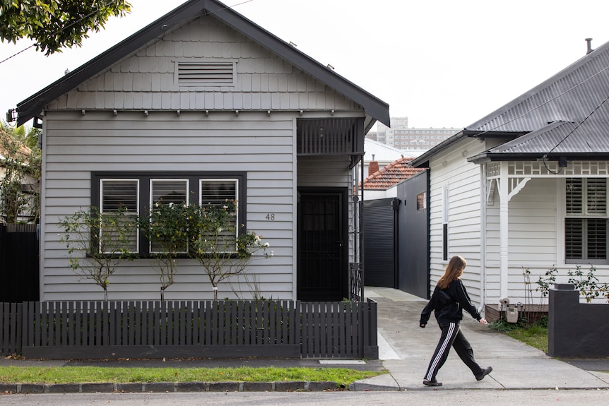 A person walks down a suburban footpath past a weatherboard house under grey skies.