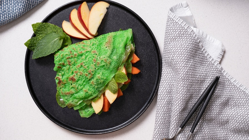 A green pancake folded into a cone on a plate with sliced apples, papaya and kaya.