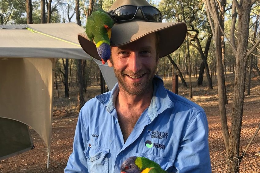 a man with a hat and blue workshirt smiles. There are rainbow lorekeets on his hat and in his hand