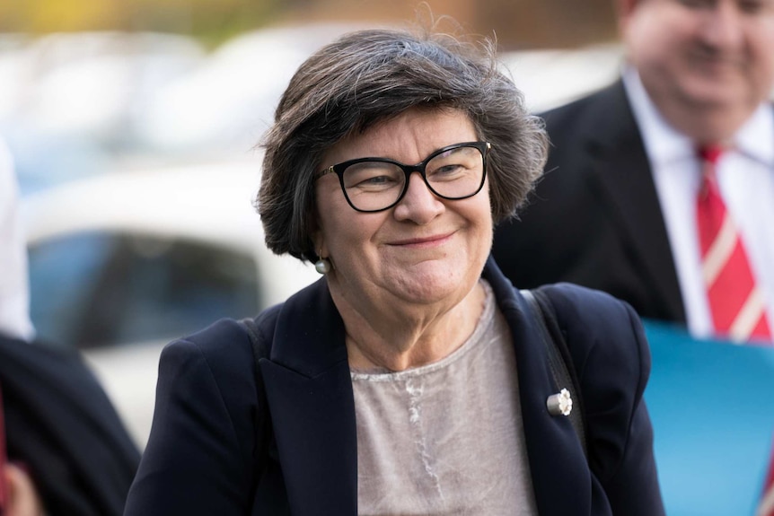 A smiling, bespectacled dark-haired woman of middle age walking down a street.