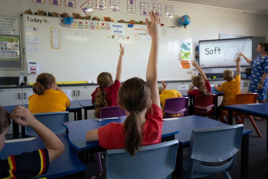 Several students raise their hands to their teacher in a primary school classroom. 