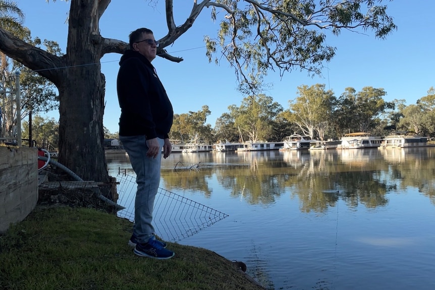 Man wears jeans, black top, sunglasses, slight paunch, standing by the river with houseboats in background. Blue waters, skies.