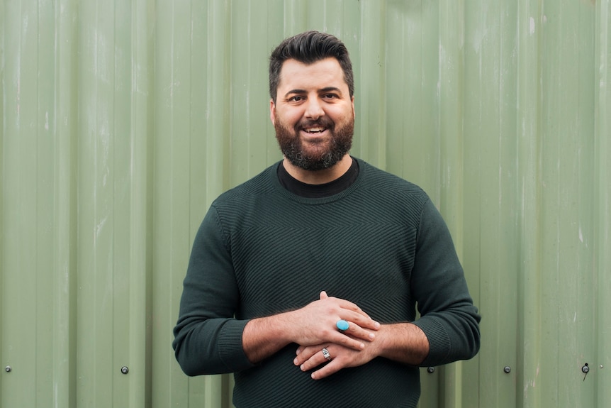 A man in a dark green shirt stands in front of a light green wall. He is smiling and wears a turquoise ring.