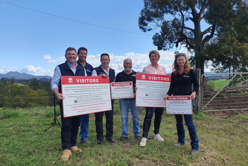 A farmer and politicians stand on a farm holding biosecurity warning signs.