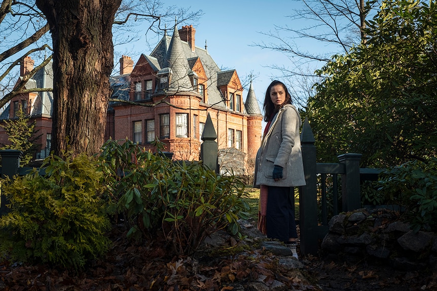 Ana de Armas stands between tree and bushes at entrance to a property featuring a large Victorian mansion on a clear blue day.