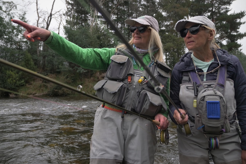 Two women holding rods and wearing lots of fly fishing equipment, standing in a river, looking into the distance.