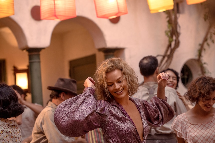 A blonde woman in her 30s, wearing a sparkling 30s-style dress, dances in a laneway, with others dancing around her