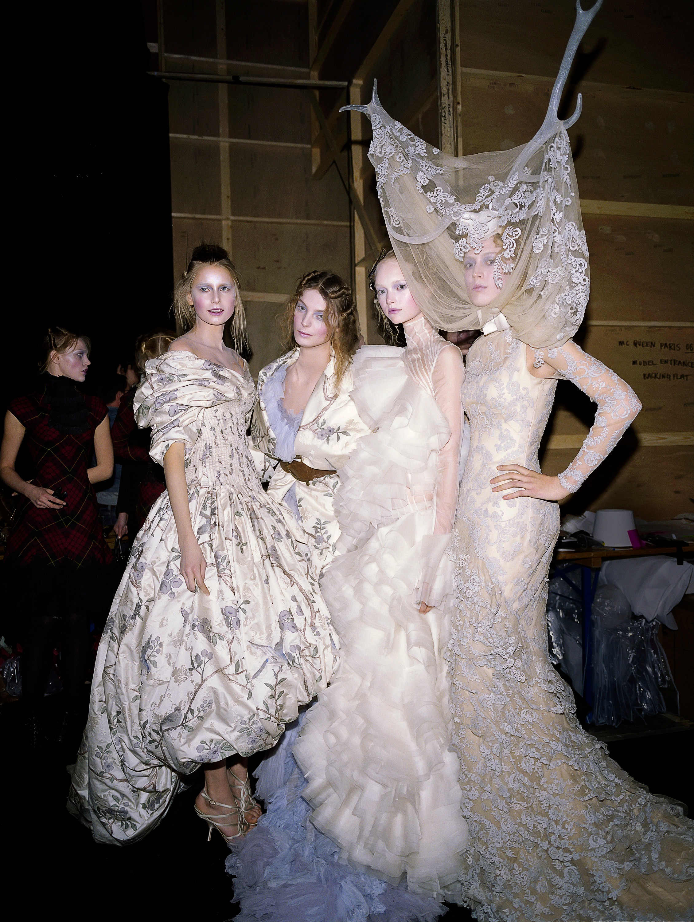 Four models pose backstage at a fashion show, all with white powdered faces and wearing extravagant white dresses