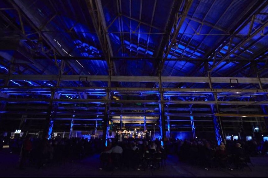 An industrial workshop at night, with an orchestra performing in front of an audience among blue lighting.