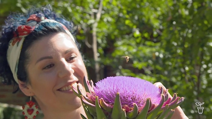 Woman smiling as she watches a bee landing on a large purple flower in a garden