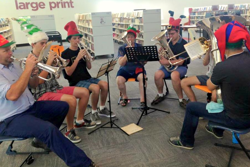 Seven people in casual clothes and Christmas hats sit in a semicircle playing brass instruments in a library.