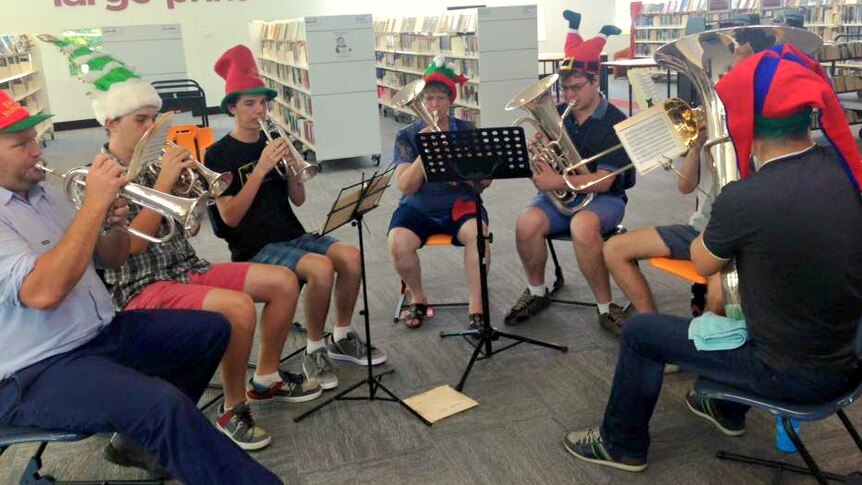 Seven people in casual clothes and Christmas hats sit in a semicircle playing brass instruments in a library.