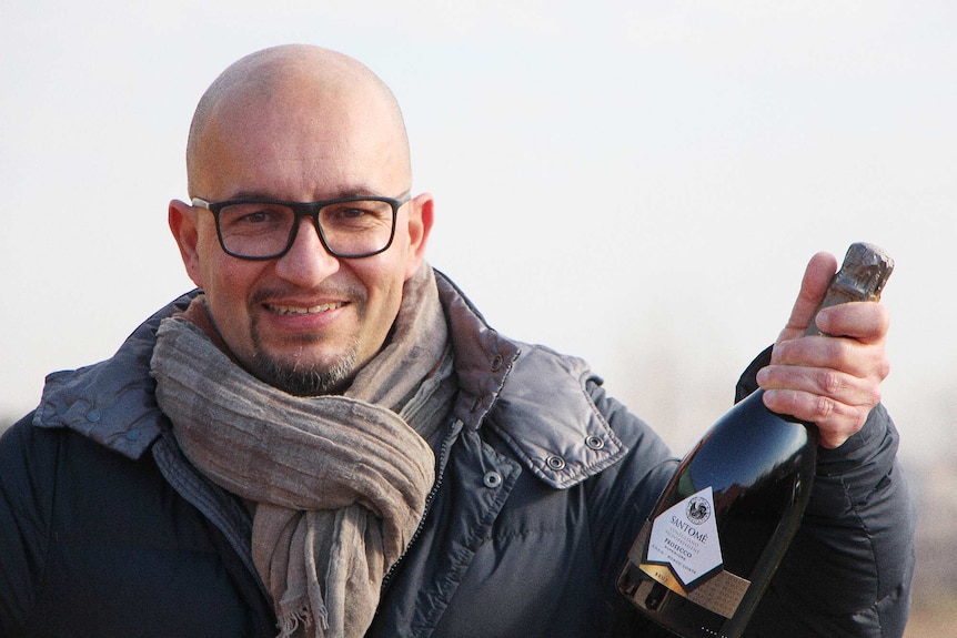William Spinazze holding a bottle of Santome Prosecco Brut in valley of vineyards, Italy.