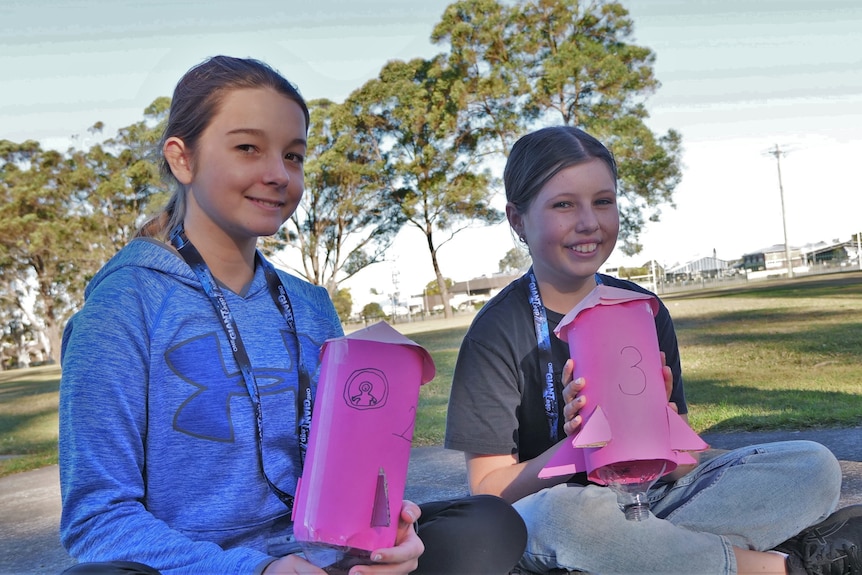 Two smiling young girls holding pink rockets with trees and grass in background.