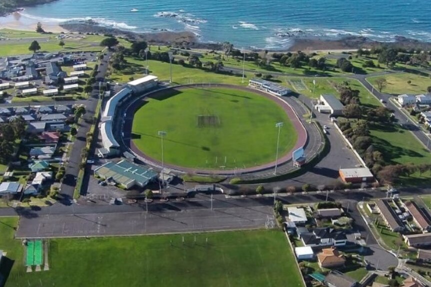 An aerial photo of an oval sporting complex with a green grounds in the middle and the beach with blue waters in the distance.