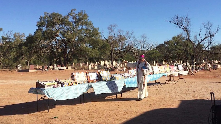 A priest in robes sprinkles holy water on colourful headstones sitting on a row of tables on red dirt at a cemetary.