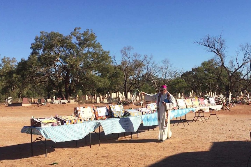 A priest in robes sprinkles holy water on colourful headstones sitting on a row of tables on red dirt at a cemetery.