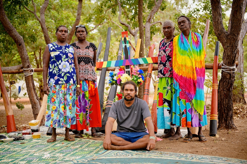 Four women standing and a man sitting in front of colorful Yidak didgeridoo. 