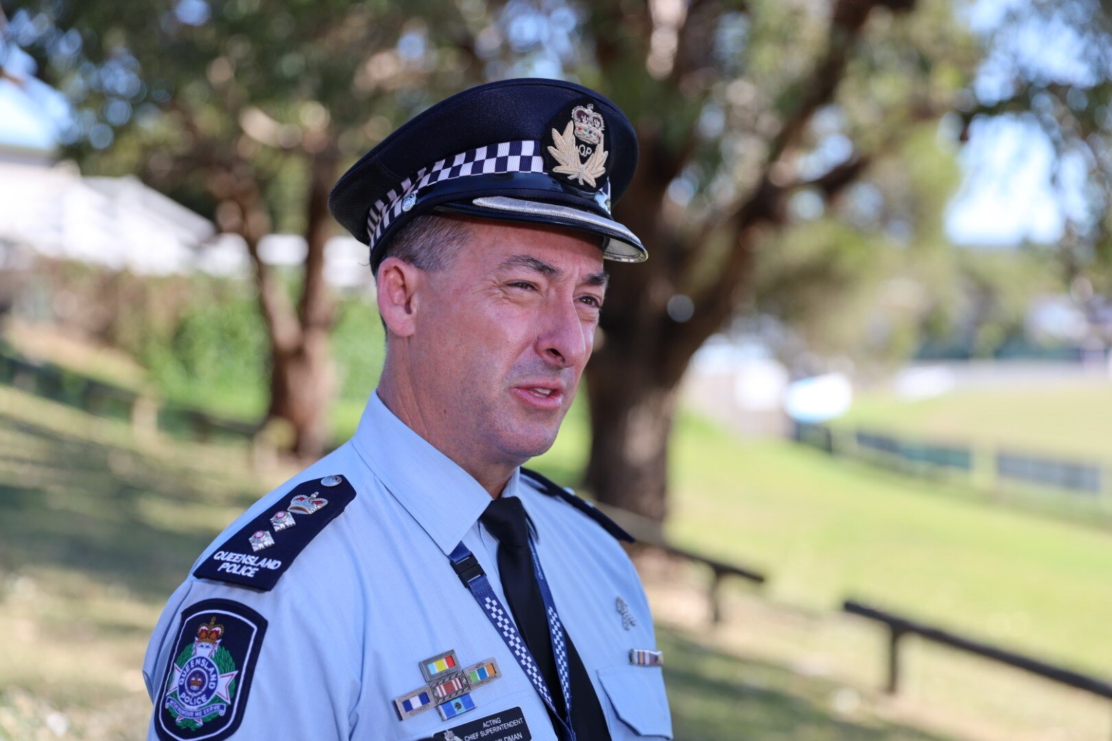 Police man wearing blue police hat speaks while standing in front of green sports field near trees