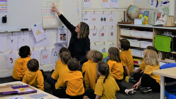 twelve students sit on the mat looking at teacher who is pointing at whiteboard.