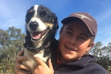 A teenager in a baseball cap and navy jumper holds up a dog.
