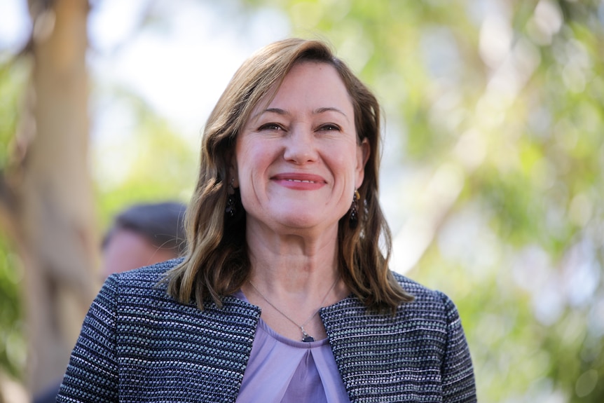 A woman stands smiling with green trees blurred out behind her.