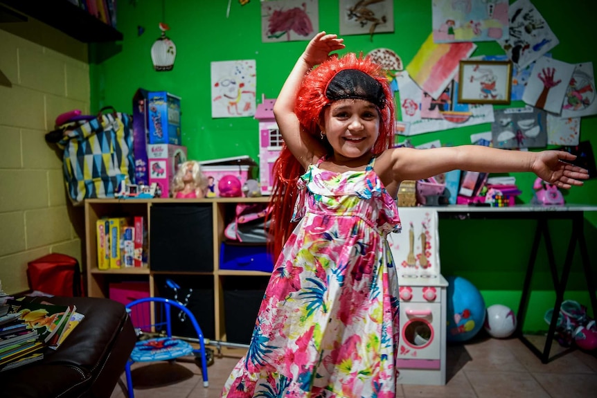 Karla De Lautour wears a wig and smiles at the camera while dancing in room with toys in the background.