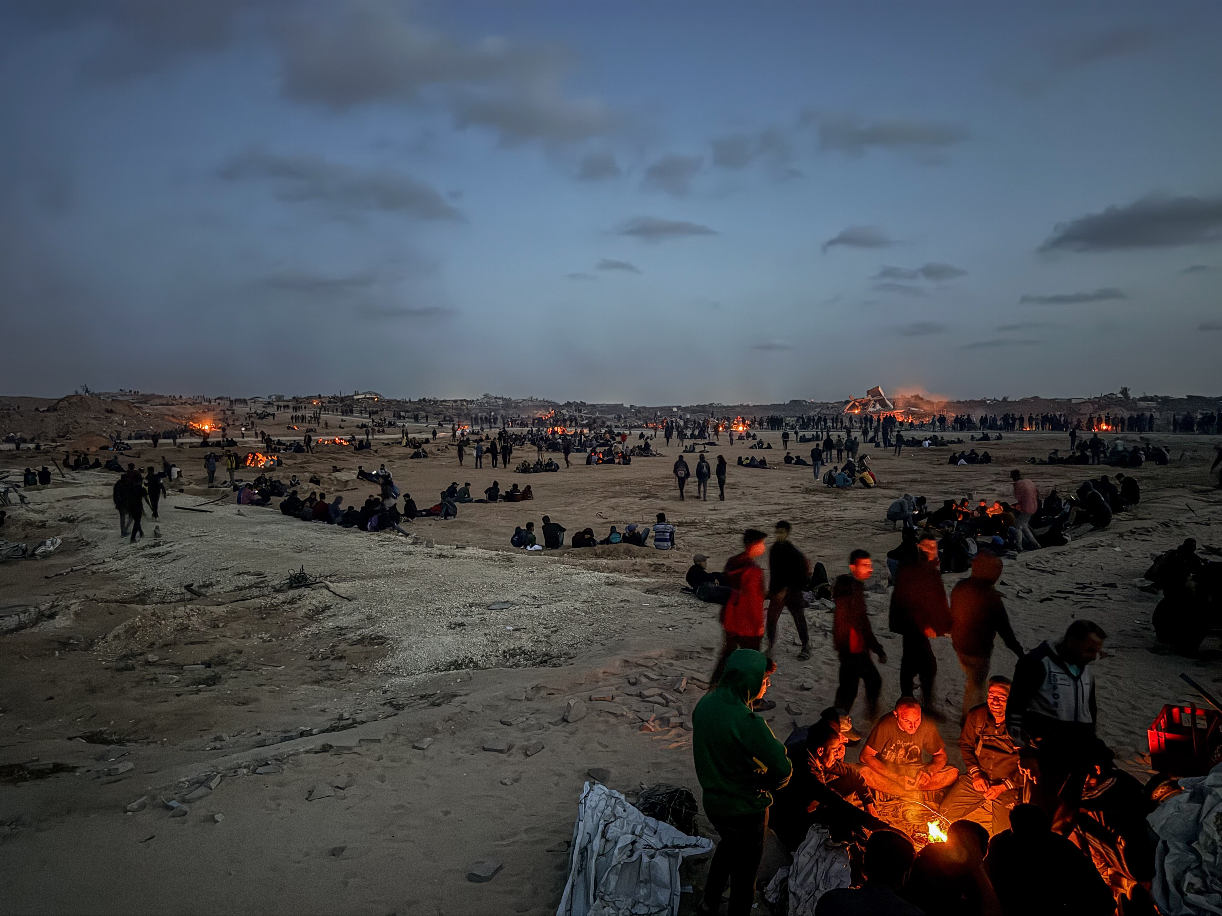 Men clusters in small groups spread across barren ground, some around orange fires, as clouds roll through the sky at dusk.
