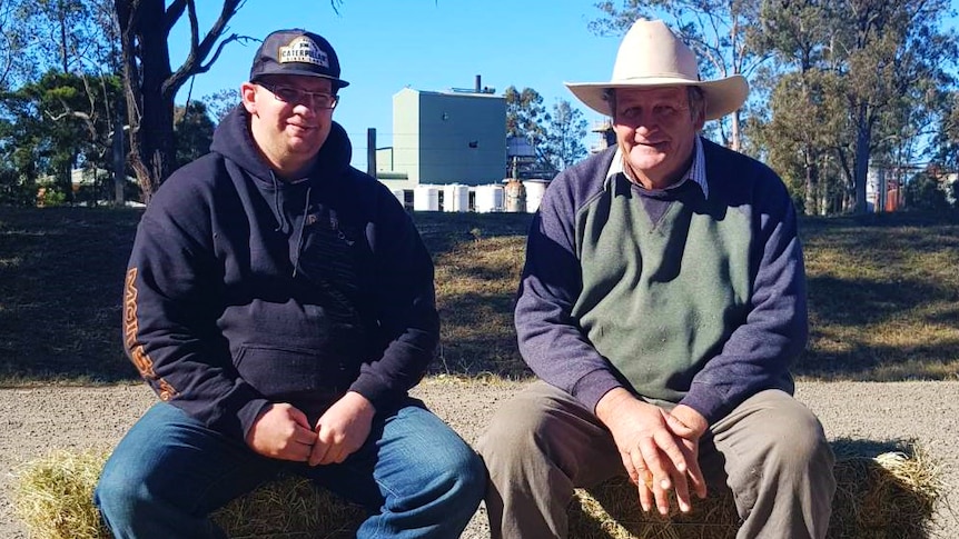 Two men sit on hay bales on gravelled ground. Both men are in jumpers and hats.