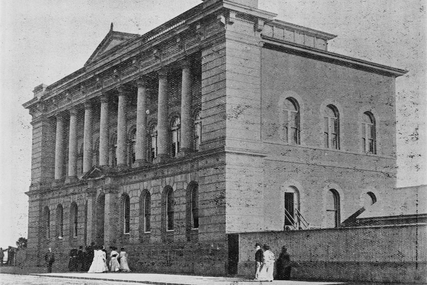 A historic picture of a large building with people walking by.
