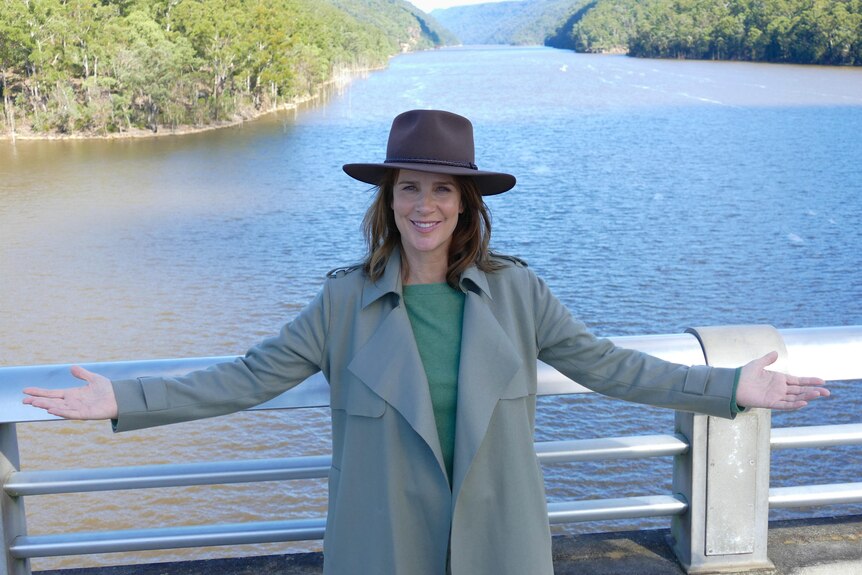 Rachel Griffiths stands on a bridge over a river with her arms spread. 