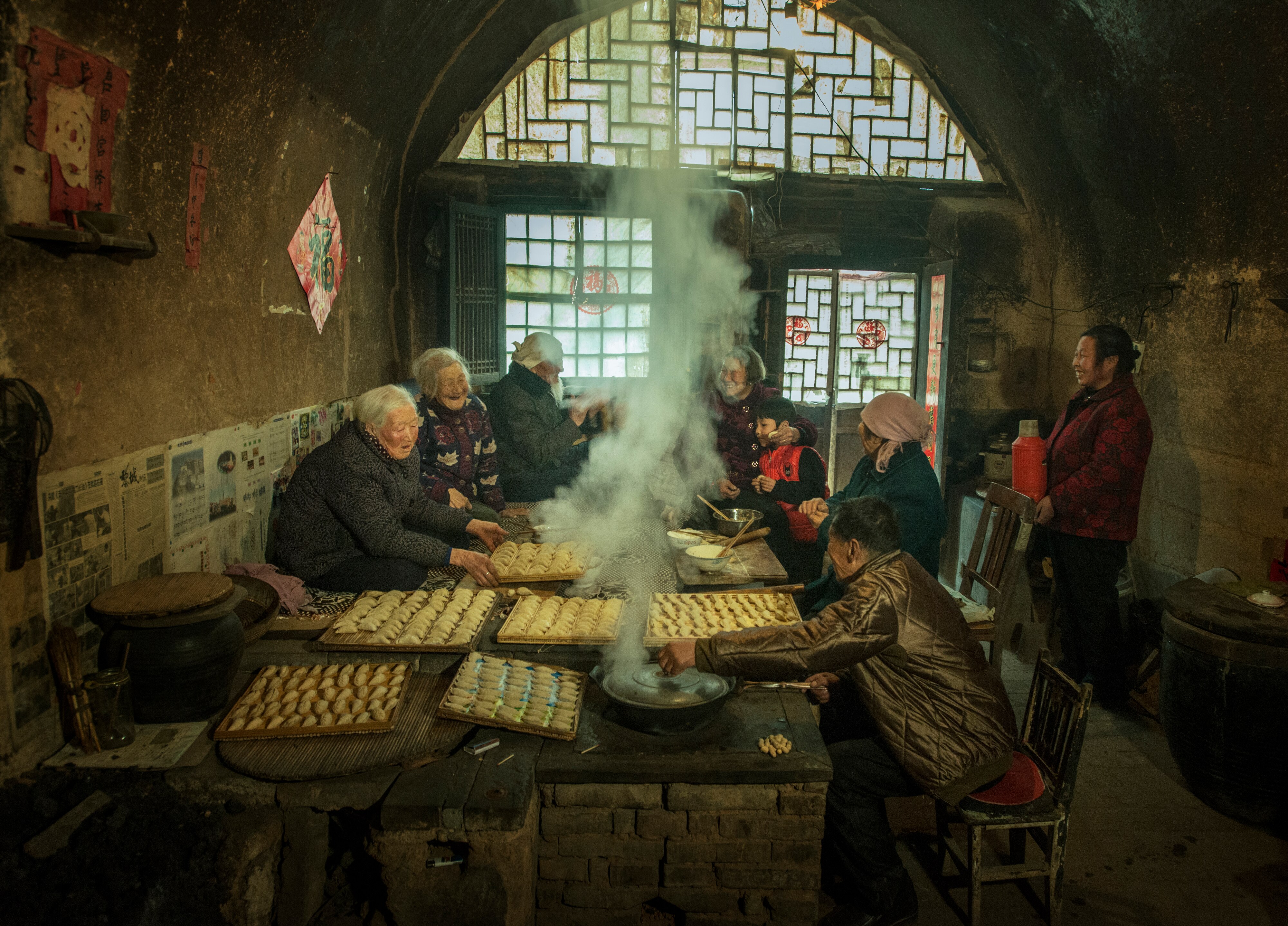 A family laughs around a table in a low-lit room. Rows of dumplings sit on the table as steam billows to the round roof