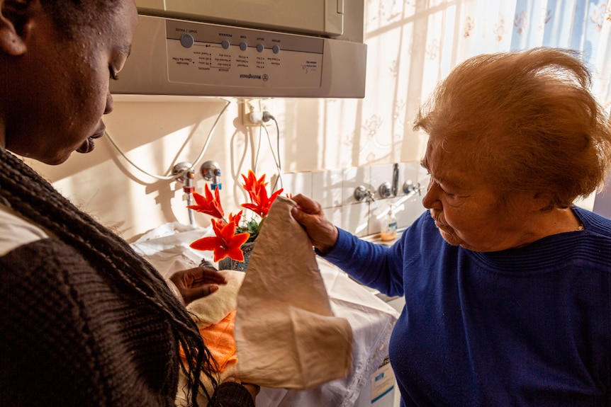 An elderly woman holding a kitchen towel above the washing machine and showing it to a younger woman.
