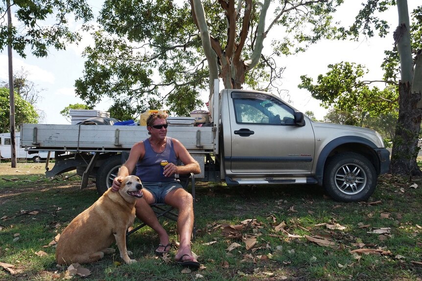 A man, with a beer in hand, sits with his dog in front of his ute near a country pub