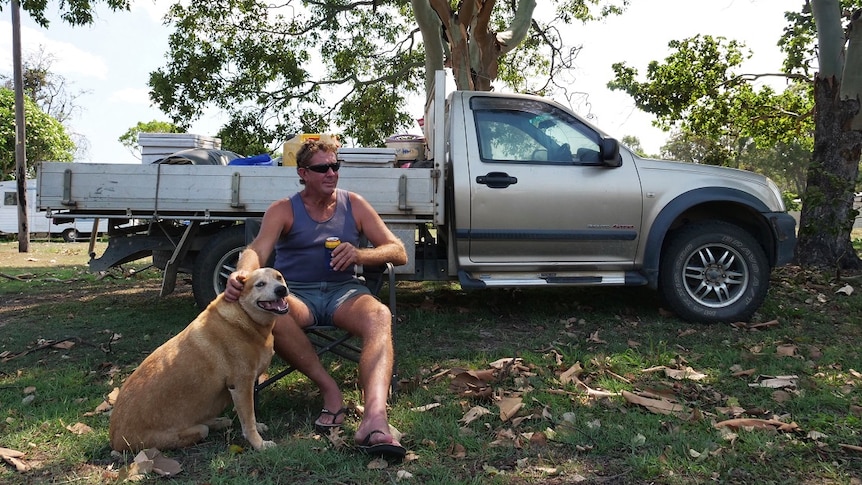 A man, with a beer in hand, sits with his dog in front of his ute near a country pub