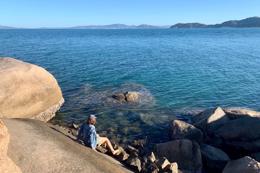 A woman sits on a rock looking out over a blue bay with mountains in the distance.