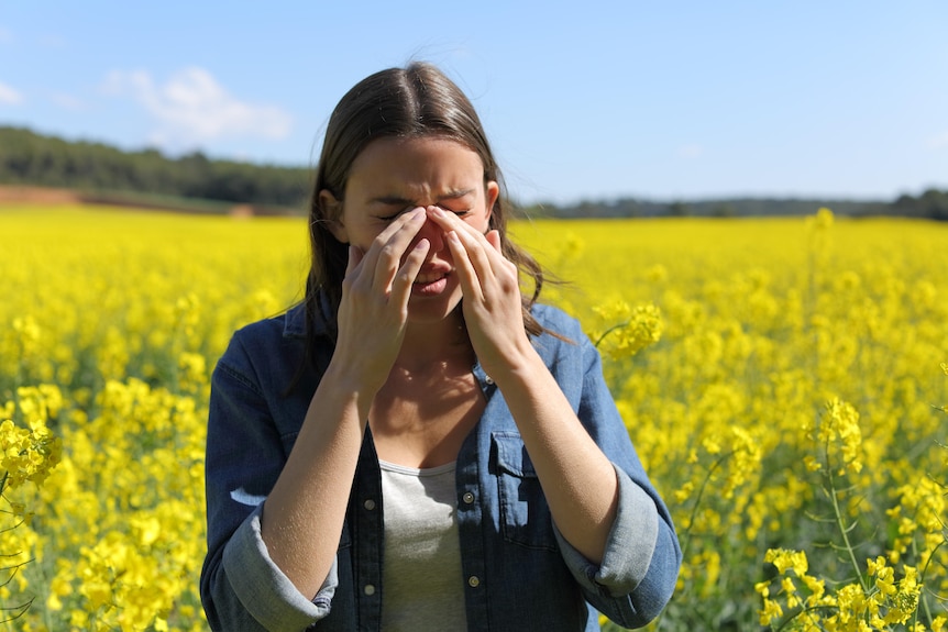A woman stands in front of a field of yellow flowers with her hands over her nose.