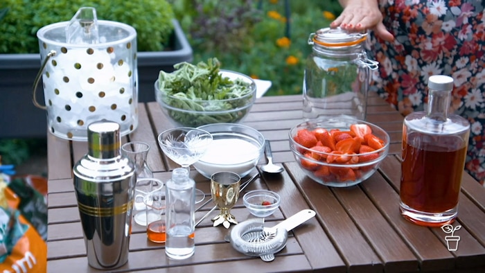 Outdoor table covered with bowls of strawberries, geranium leaves and sugar and cocktail making equipment
